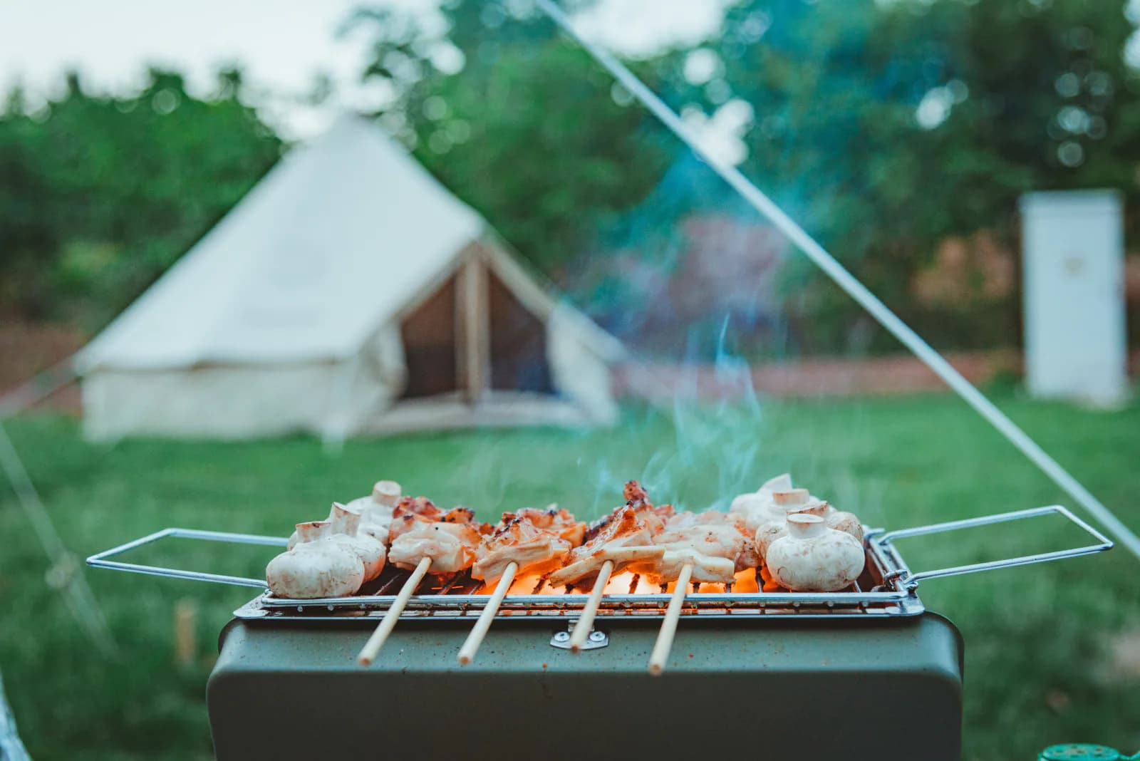 Skewers cooking on a portable grill in front of a white canvas tent at camp