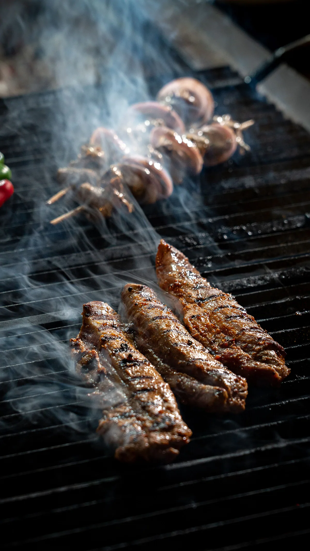 Meat skewers being moved across a smoky grill grate with tongs