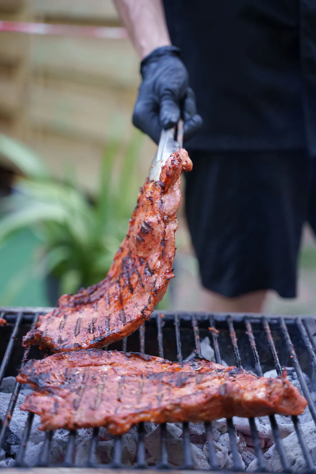 A cook lifting a rack of grilled meat with tongs above a charcoal grate